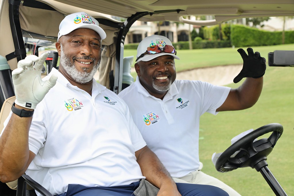Two men in white golf shirts and caps sit smiling in a golf cart on a green golf course, waving at the camera. South Florida Business & Wealth