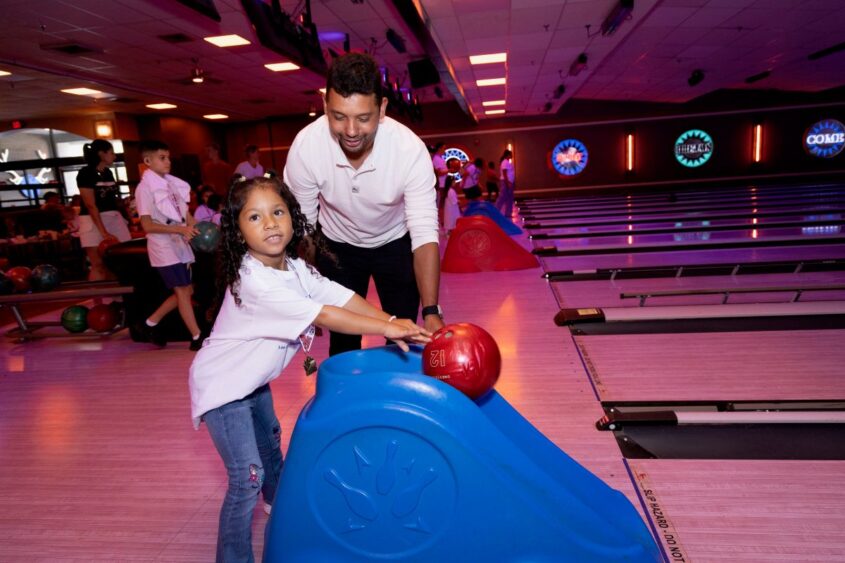 A young girl uses a bowling ramp with help from an adult man at a bowling alley. Other people bowl in the background under colorful lights. South Florida Business & Wealth