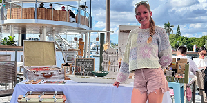 A woman smiles while standing by a table displaying handmade crafts and jewelry at an outdoor market with boats and palm trees in the background. South Florida Business & Wealth
