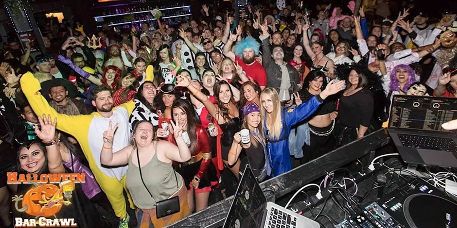 A large group of people in costumes pose and smile at a lively Halloween bar crawl event, with DJs and party lights in the background. South Florida Business & Wealth