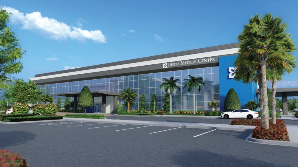 Modern medical center with large glass windows, a “Jupiter Medical Center” sign, palm trees, landscaped greenery, parked cars, and a clear sky with scattered clouds. South Florida Business & Wealth