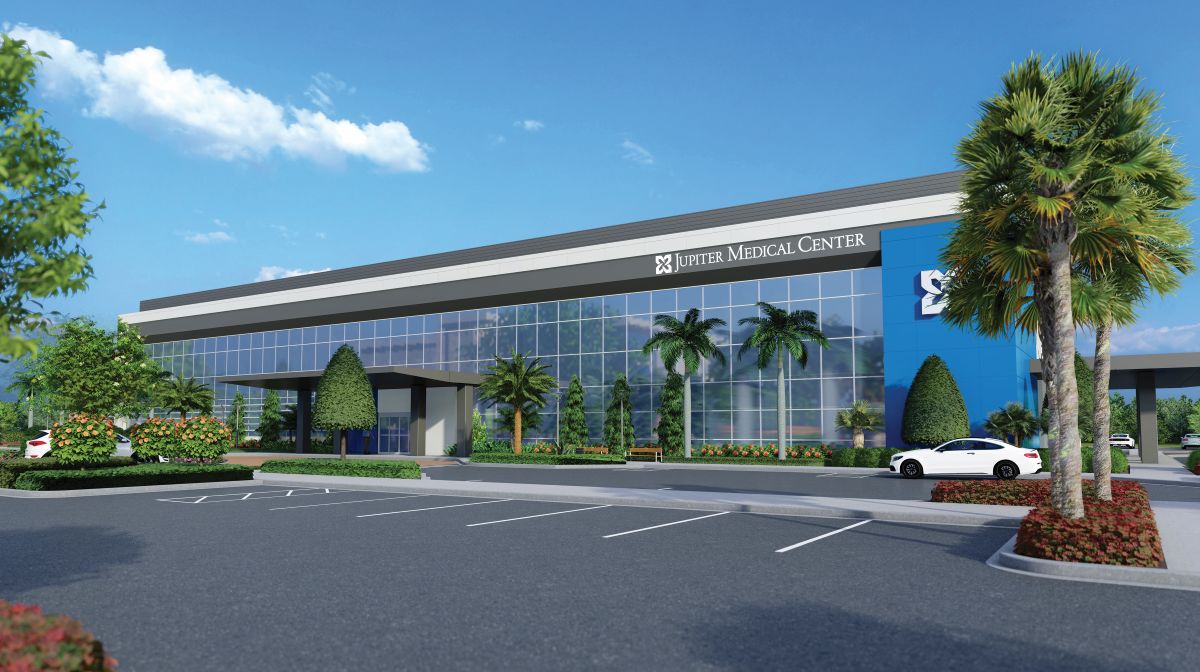 Modern medical center with large glass windows, a “Jupiter Medical Center” sign, palm trees, landscaped greenery, parked cars, and a clear sky with scattered clouds. South Florida Business & Wealth