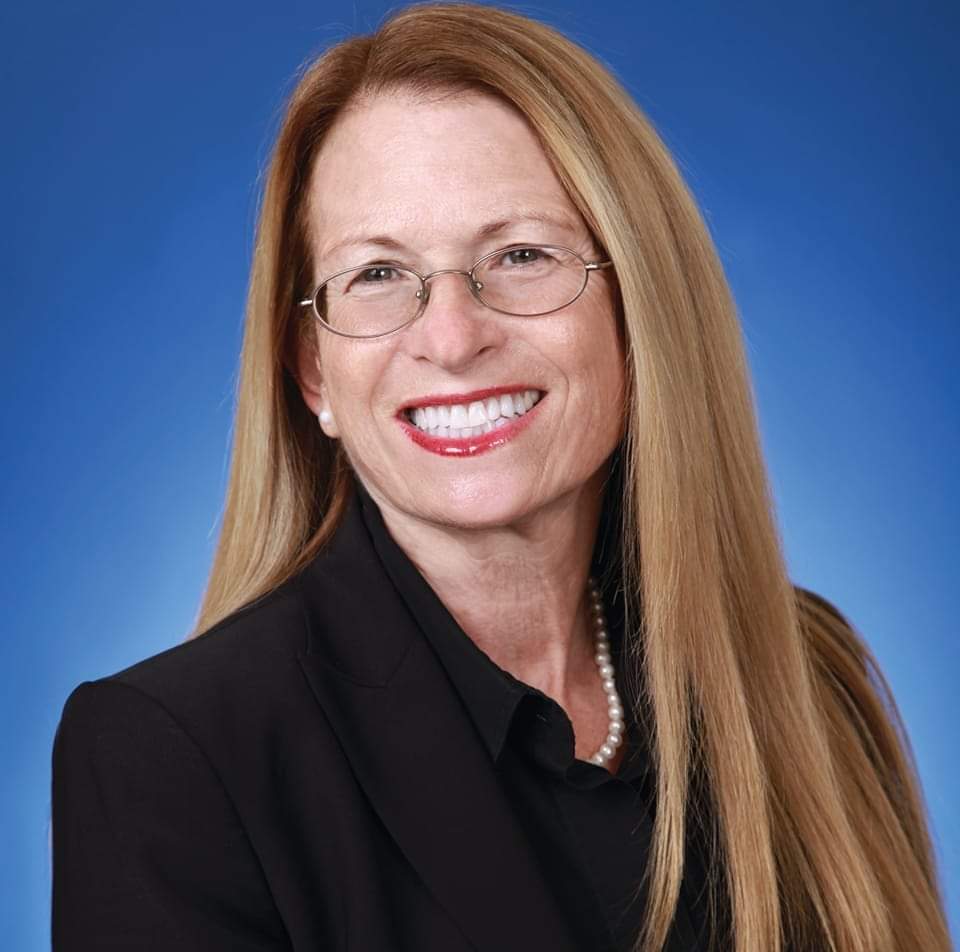 Smiling woman with long straight blonde hair, wearing glasses, a black blazer, and a pearl necklace, posed in front of a solid blue background. South Florida Business & Wealth