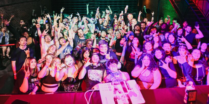 A large, energetic crowd faces the camera at an indoor concert or event, many smiling, cheering, and making hand gestures, under bright colorful stage lights. South Florida Business & Wealth
