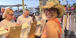 A smiling woman in a hat and sunglasses stands by a jewelry display at an outdoor market, with other shoppers browsing nearby and string lights overhead. South Florida Business & Wealth