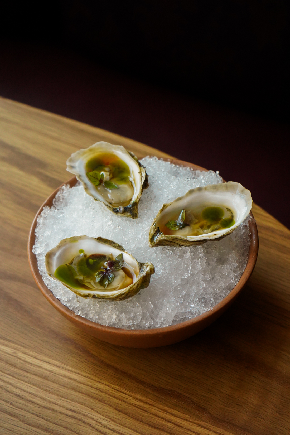 Three half-shell oysters with green garnish are served on a bed of crushed ice in a round wooden bowl on a wooden table. South Florida Business & Wealth