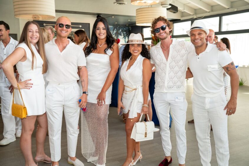 Six people stand together indoors, smiling and dressed in stylish white outfits. Three women and three men pose for the camera, some wearing sunglasses and hats. The background shows modern decor and soft natural light. South Florida Business & Wealth