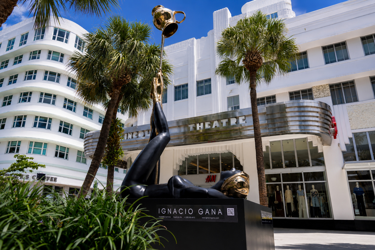 A black and gold reclining sculpture by Ignacio Gana sits among palm trees in front of a white Art Deco building and theater on a sunny day. South Florida Business & Wealth