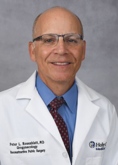 A smiling man wearing glasses, a white lab coat with a Holy Cross Medical Group logo, a blue shirt, and a red patterned tie, standing in front of a gray background. South Florida Business & Wealth