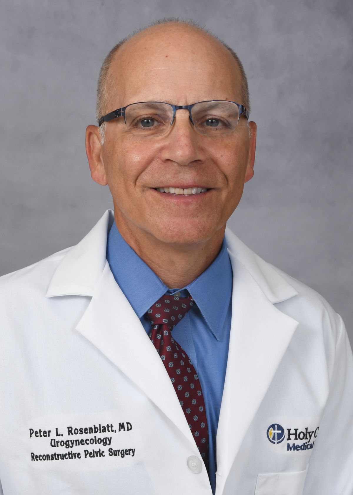 A smiling man wearing glasses, a white lab coat with a Holy Cross Medical Group logo, a blue shirt, and a red patterned tie, standing in front of a gray background. South Florida Business & Wealth