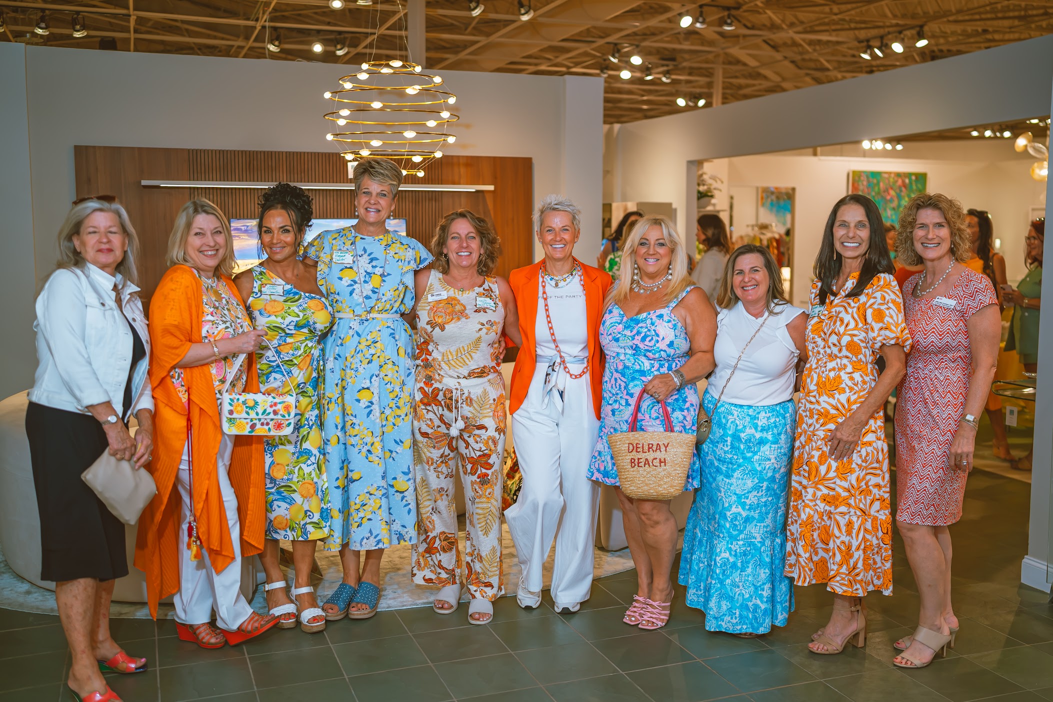 Ten women stand side by side indoors, smiling, dressed in colorful orange, white, and blue summer outfits. Modern lighting and artwork are visible in the background. South Florida Business & Wealth