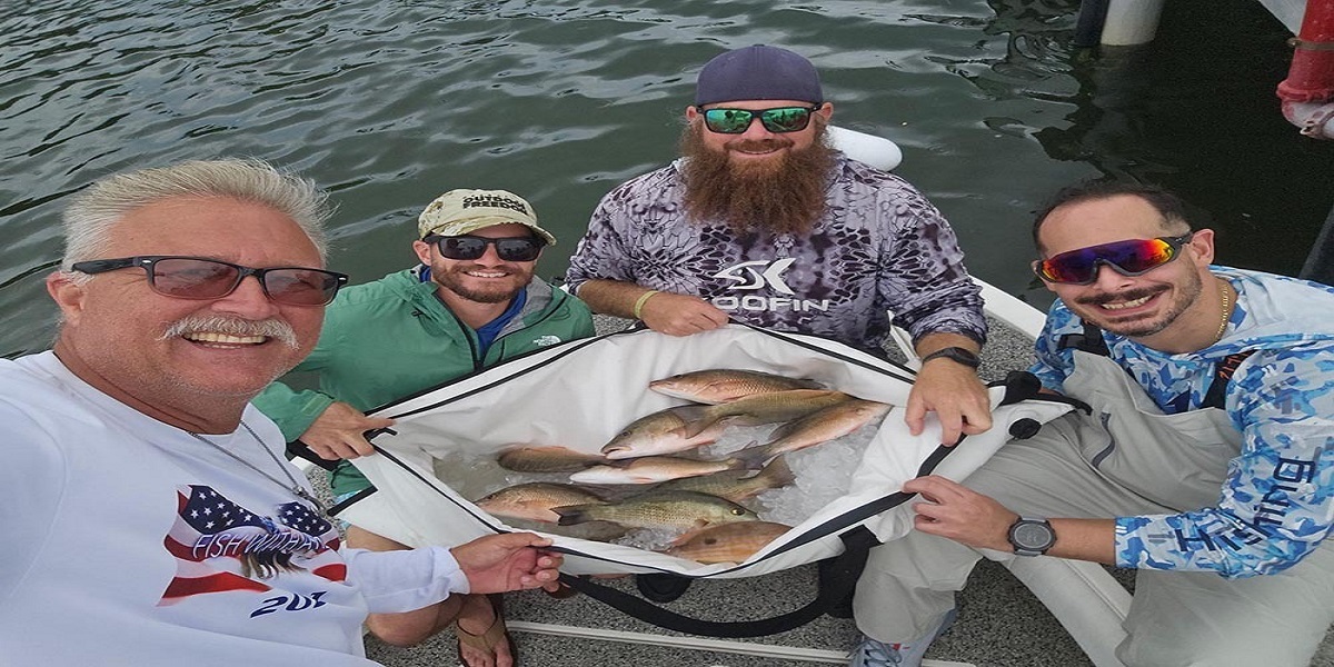 Four smiling men on a boat pose with a large cooler full of freshly caught fish, with water visible in the background. South Florida Business & Wealth
