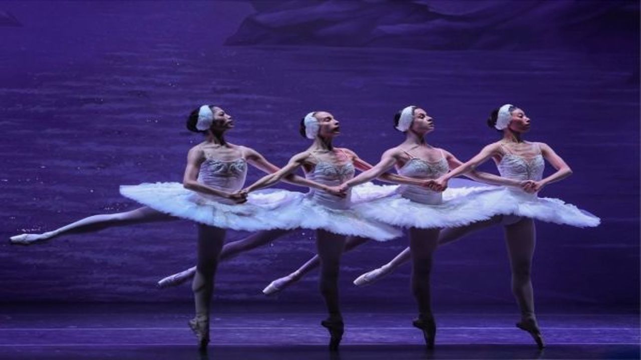 Four ballerinas in white tutus perform in unison on stage, standing on tiptoe with arms interlinked, against a purple-lit backdrop suggesting a lake scene. South Florida Business & Wealth