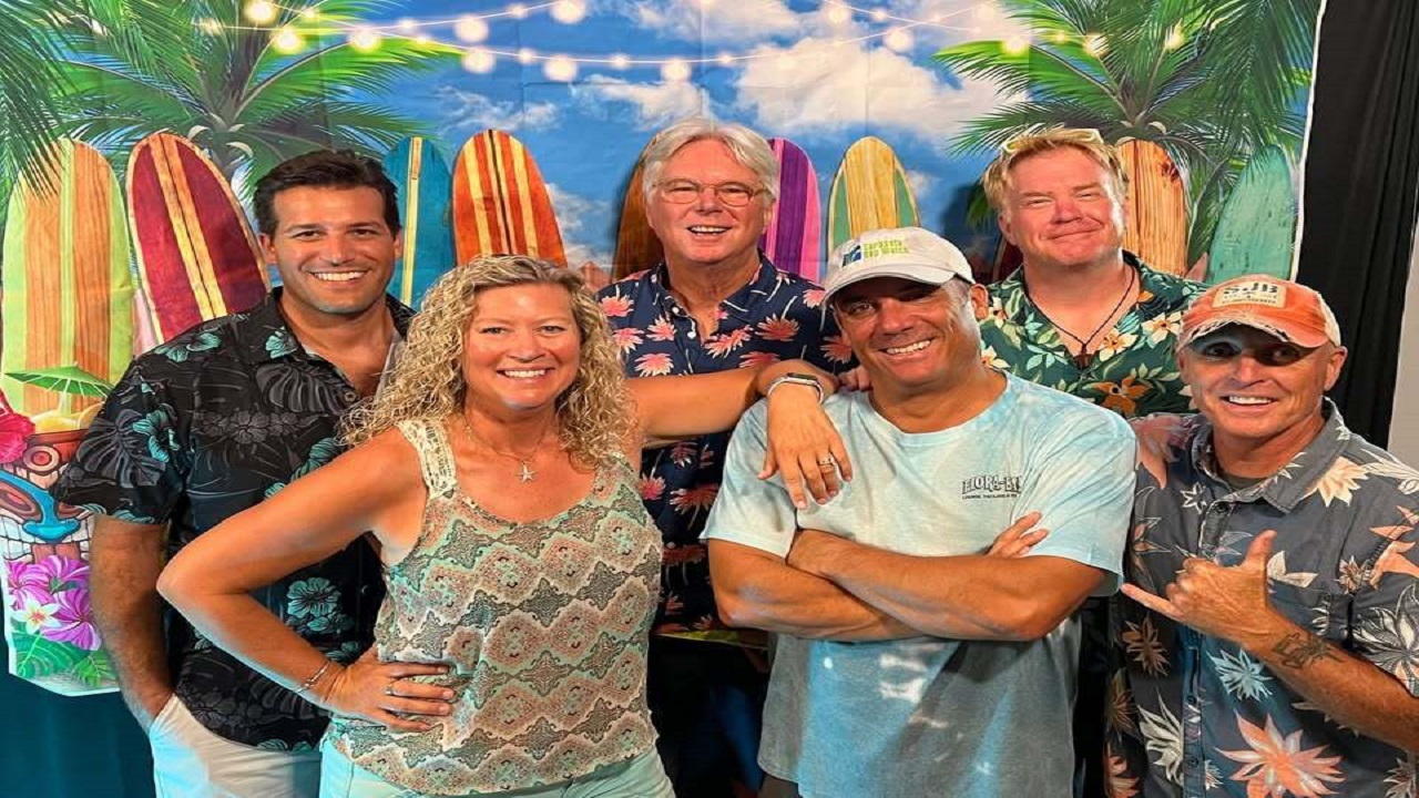 Six smiling adults in tropical shirts pose in front of a beach-themed backdrop with palm trees and surfboards. String lights hang overhead, creating a festive, summery atmosphere. South Florida Business & Wealth