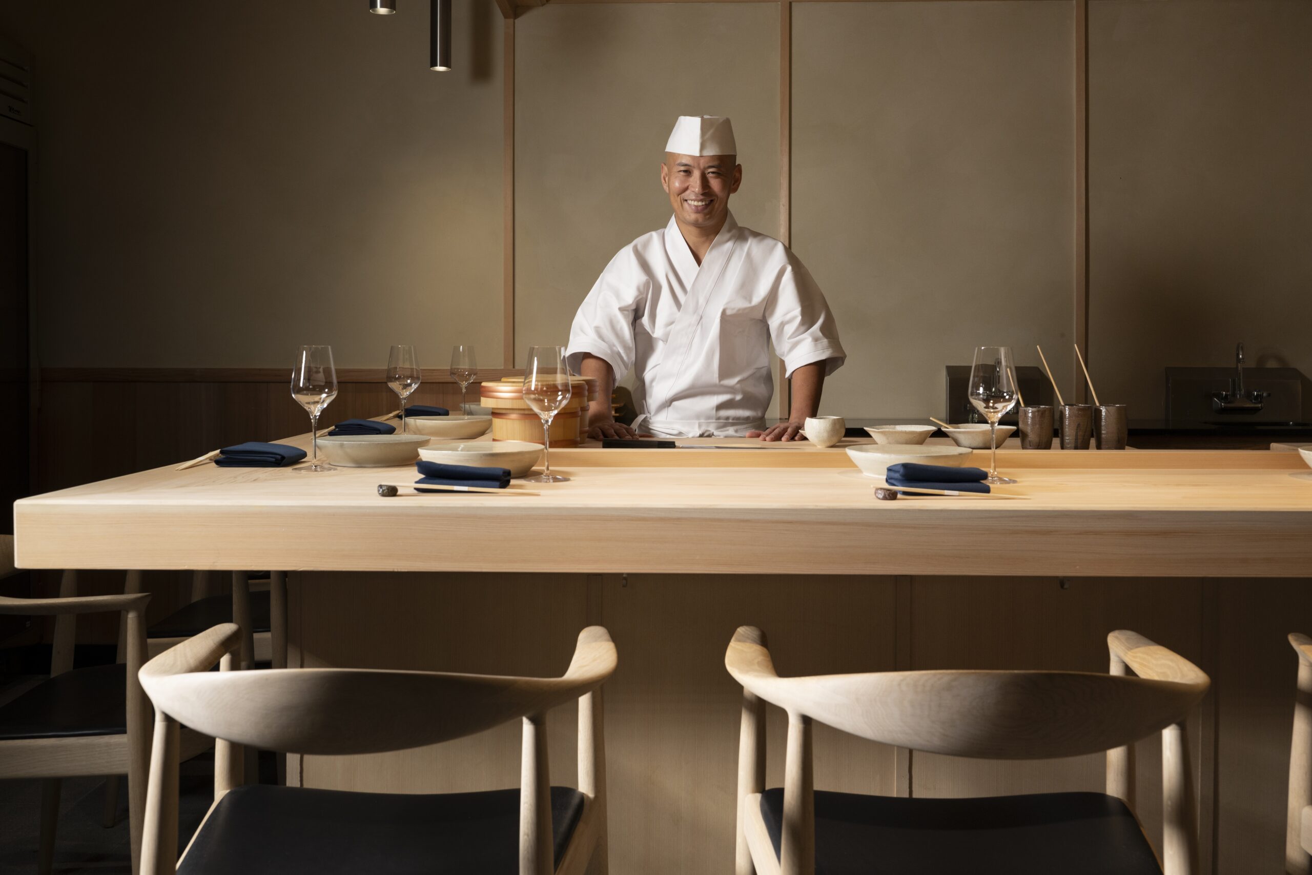 A chef in traditional attire stands smiling behind a wooden sushi counter, set with plates, glasses, and utensils in a minimalist, softly lit restaurant. South Florida Business & Wealth