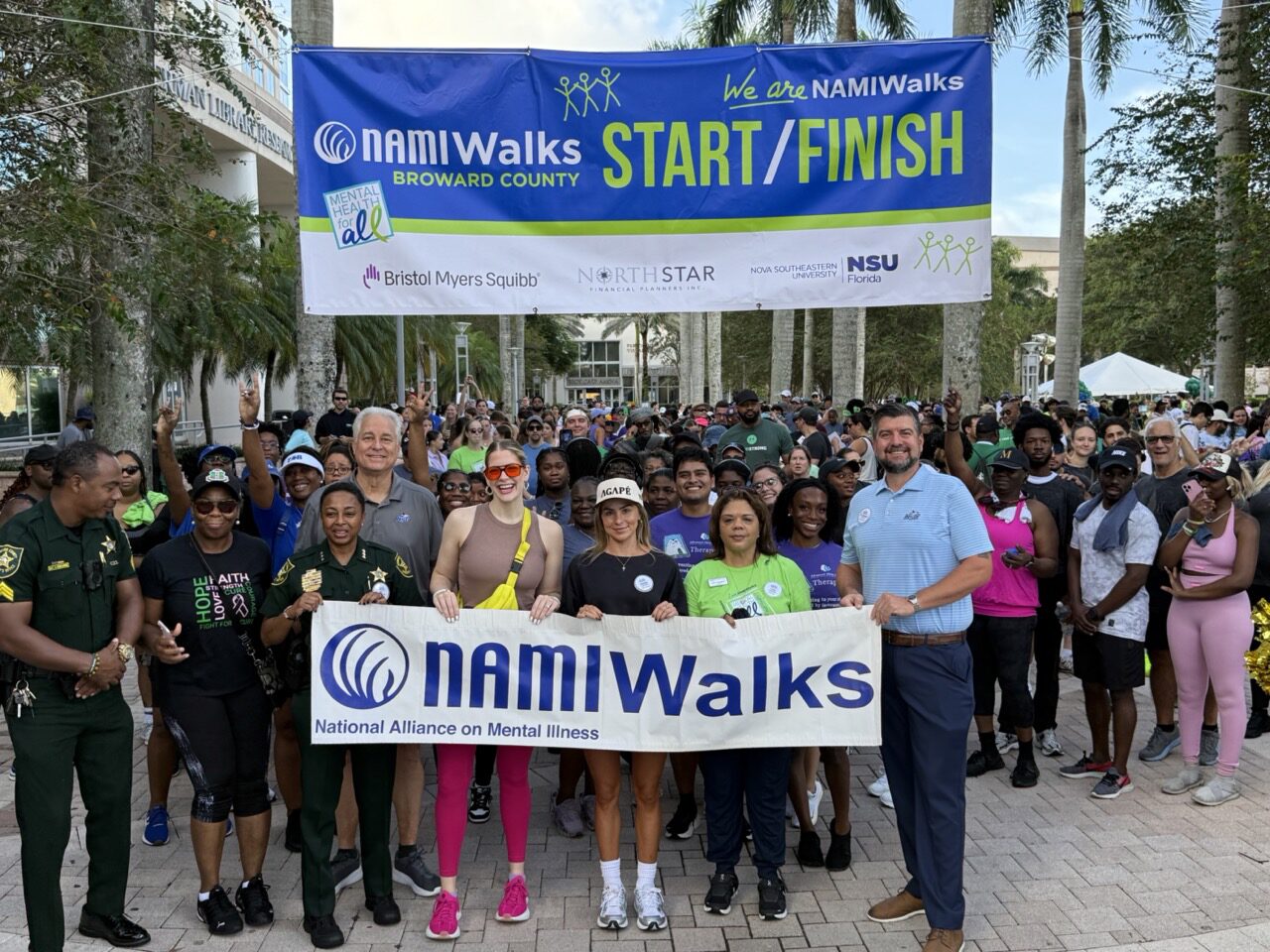A large group of people at the NAMIWalks event stand under a “Start/Finish” banner, smiling and holding a NAMIWalks sign. Palm trees and event booths are visible in the background. South Florida Business & Wealth