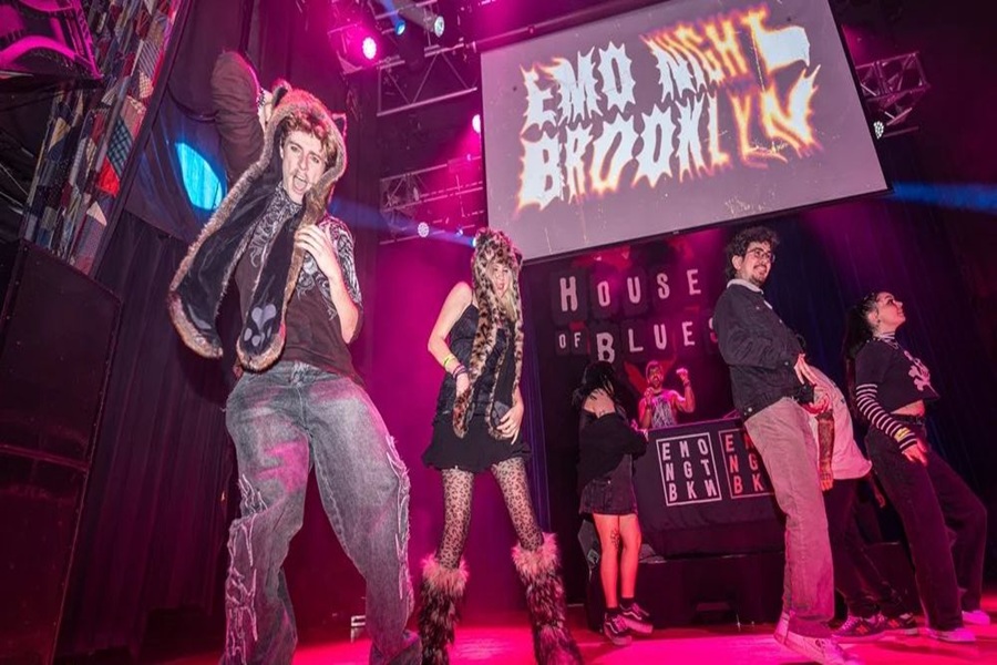 People dance on a stage under colorful lights at an event called “Emo Night Brooklyn” at the House of Blues. A large sign and a DJ booth are visible in the background. The mood is lively and energetic. South Florida Business & Wealth