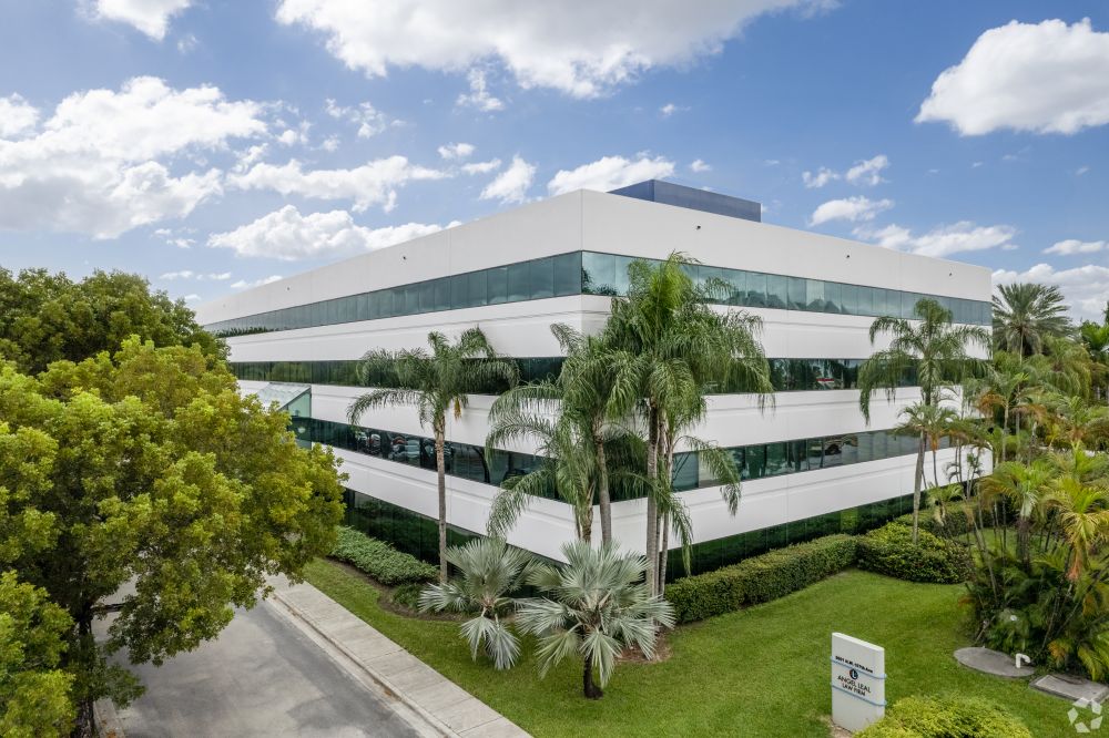 A modern white office building with mirrored green windows, surrounded by palm trees and greenery, under a partly cloudy blue sky. South Florida Business & Wealth