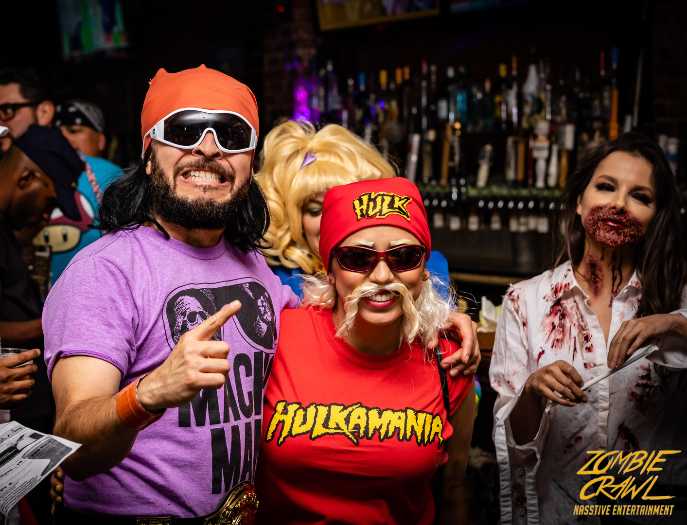 Three people in costumes: one in a purple “Macho Man” shirt, one in a red “Hulkamania” outfit, and one as a zombie with fake blood, smiling at a bar during a themed event. “Zombie Crawl” is written in the corner. South Florida Business & Wealth