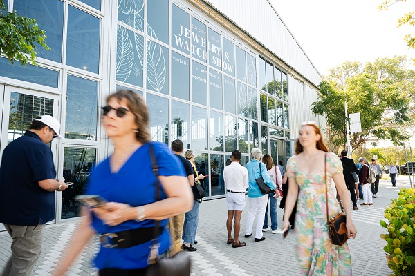 People stand in line outside a glass building with “Jewelry & Watch Show” signage. Some are walking, others are on their phones. It’s sunny, and trees line the walkway. South Florida Business & Wealth