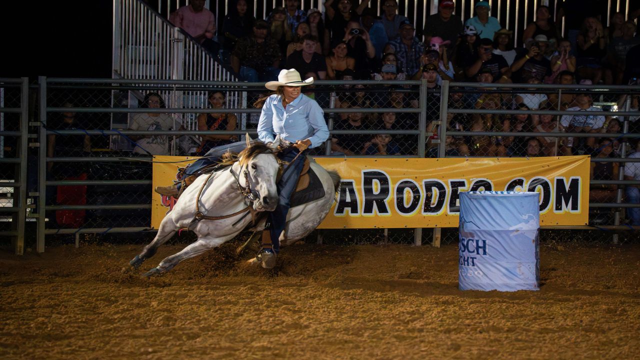 A rodeo rider wearing a cowboy hat guides a white horse around a blue barrel at night, with a cheering crowd and a yellow banner in the background. South Florida Business & Wealth