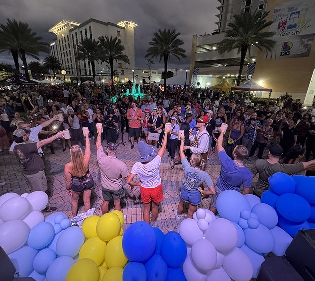 A large crowd gathers outdoors at night, raising drinks together. Blue and yellow balloons decorate the front. Palm trees and tall buildings are in the background under a cloudy sky. South Florida Business & Wealth