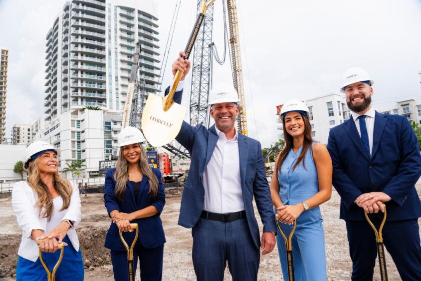 Five people in business attire and hard hats stand at a construction site, smiling. The man in the center holds a golden shovel up. Tall buildings and construction equipment are visible in the background. South Florida Business & Wealth