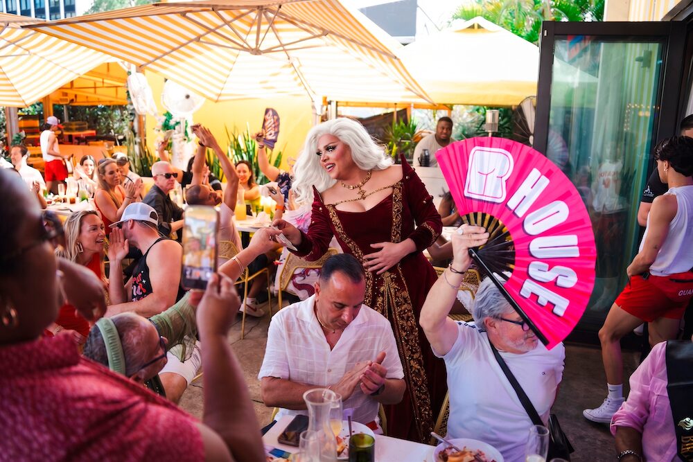 A drag queen in a red dress interacts with a cheering crowd at an outdoor brunch. People smile, clap, and hold up phones. One person holds a large pink “HOUSE” fan. Yellow umbrellas and greenery decorate the lively scene. South Florida Business & Wealth