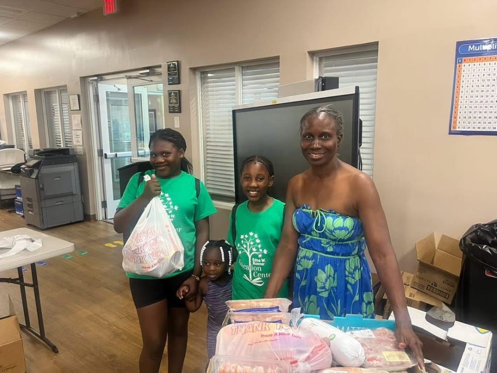 Three girls and a woman stand in a room with food items on a table. Two girls wear green shirts, the smallest girl wears purple, and the smiling woman wears a blue floral dress. All are looking at the camera. South Florida Business & Wealth