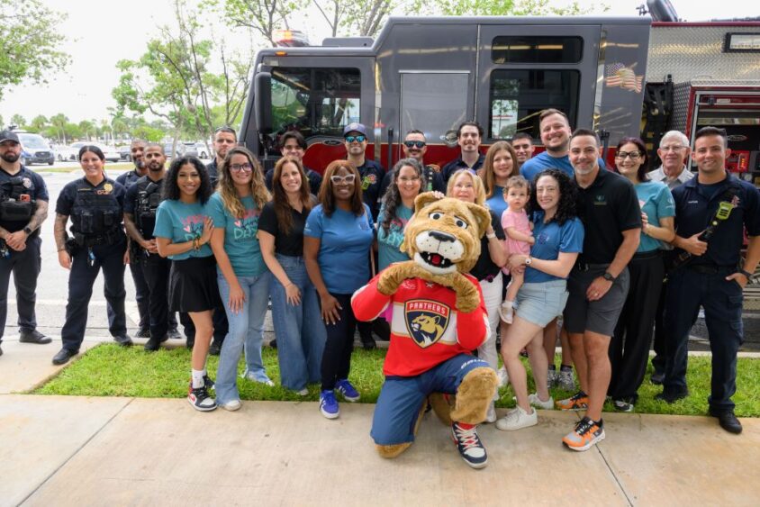 A group of people, police officers, firefighters, and a person in a panther mascot costume pose and smile together in front of a fire truck on a sunny day. South Florida Business & Wealth