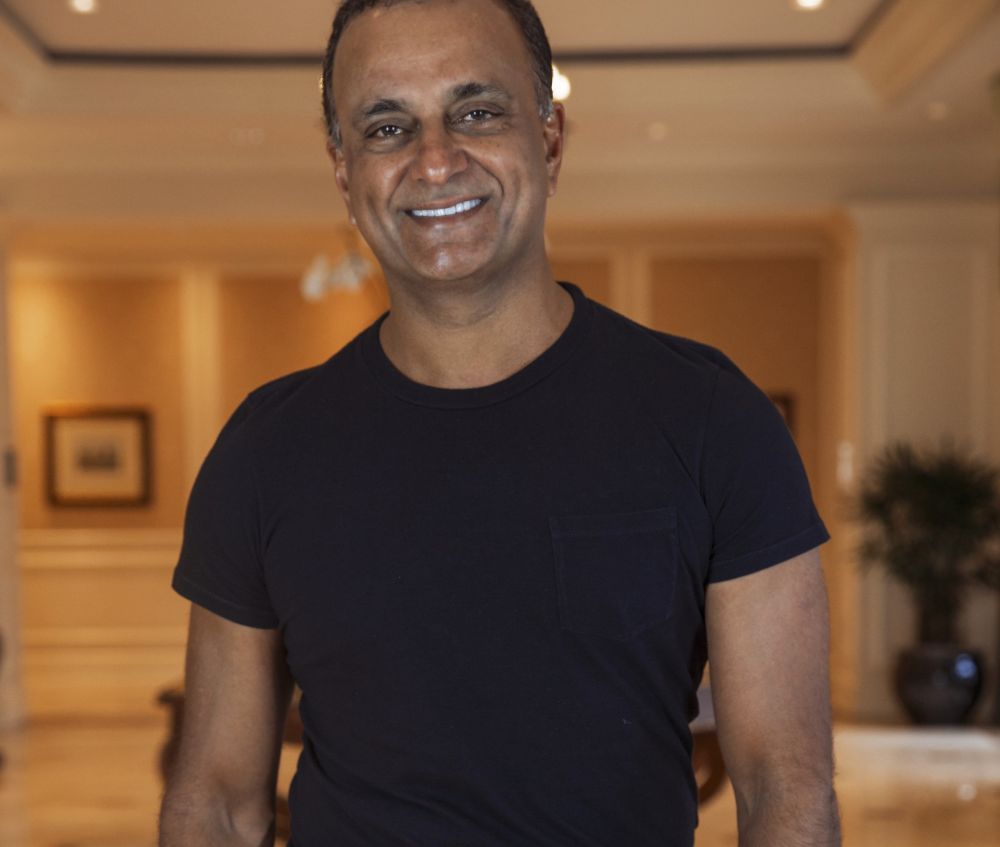 A man in a black t-shirt stands indoors, smiling at the camera. The background features warm lighting, framed art on the wall, and blurred furnishings in a spacious room. South Florida Business & Wealth