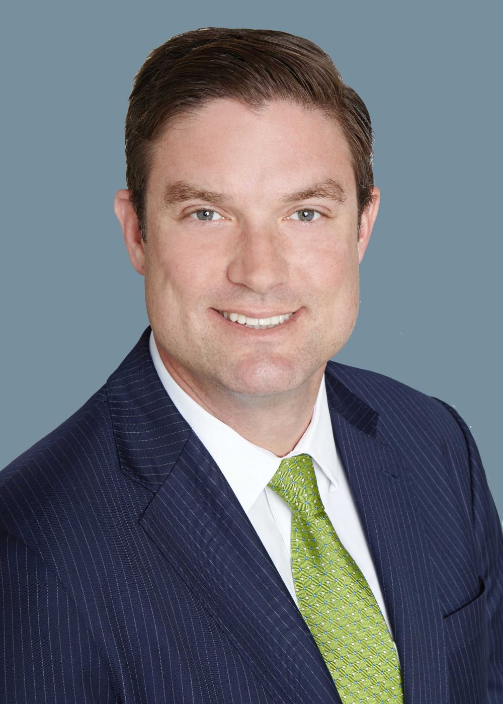 A man in a dark pinstripe suit, white shirt, and green tie smiles at the camera against a plain grey background. South Florida Business & Wealth