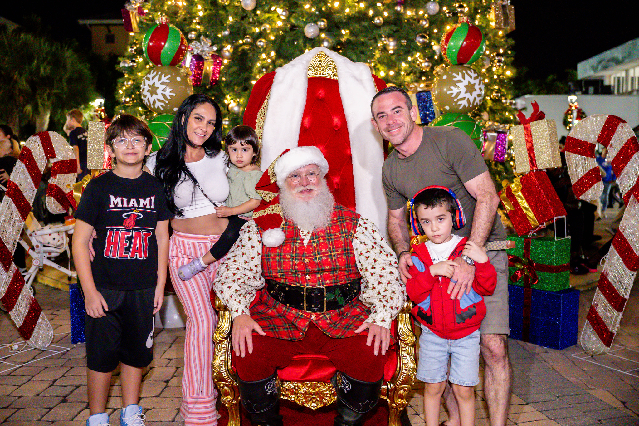 A family with three children poses with Santa Claus in front of a large decorated Christmas tree and festive ornaments, all smiling for the camera. South Florida Business & Wealth