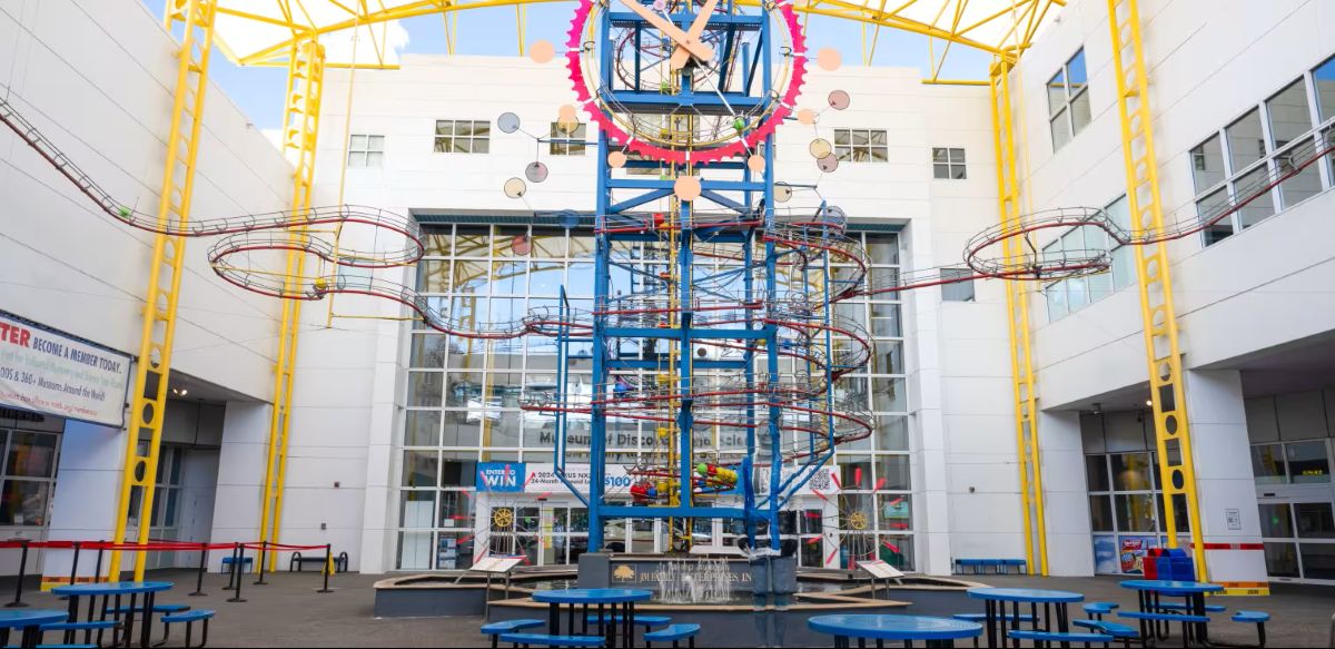 A large kinetic sculpture with winding tracks stands in a bright atrium, surrounded by blue round tables and yellow beams, with a white building and glass windows in the background. South Florida Business & Wealth