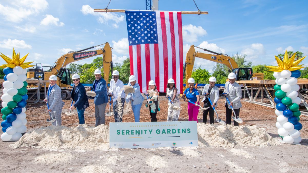 Nine people in hard hats and formal attire take part in a groundbreaking ceremony with shovels, standing before a banner reading “Serenity Gardens”; an American flag and construction equipment are in the background. South Florida Business & Wealth