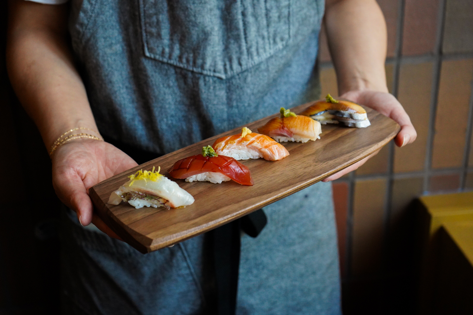 A person in a gray apron holds a wooden board with five pieces of assorted sushi, each garnished with toppings, against a tiled background. South Florida Business & Wealth