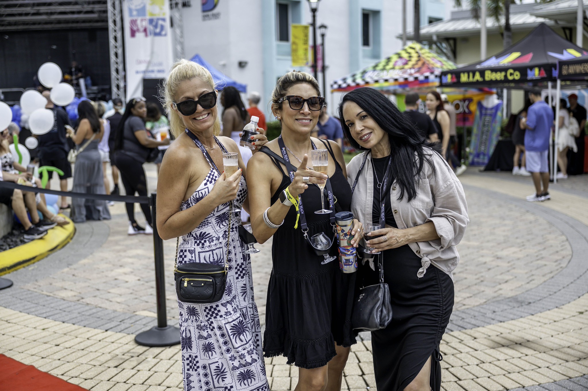 Three women stand smiling on a brick street during an outdoor event, each holding a drink. People, tents, and colorful booths are visible in the background. The women wear sunglasses and summer dresses. South Florida Business & Wealth