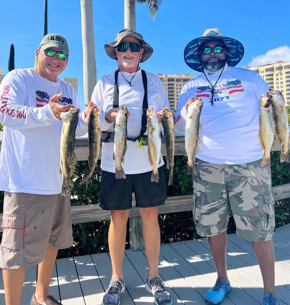 Three smiling men in hats and sunglasses hold several caught fish while standing on a dock. They wear matching white shirts and stand in front of palm trees and buildings under a clear blue sky. South Florida Business & Wealth