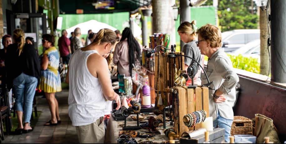People browse handmade jewelry and crafts displayed on outdoor tables at a busy market. Some are shopping, while others chat under a covered walkway lined with vendors and colorful items. Greenery is visible in the background. South Florida Business & Wealth