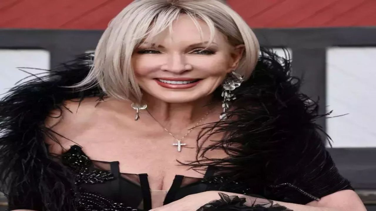 Smiling woman with short blonde hair wearing a black feathered outfit, dangling earrings, and a cross necklace, standing in front of a building with red and white panels. South Florida Business & Wealth