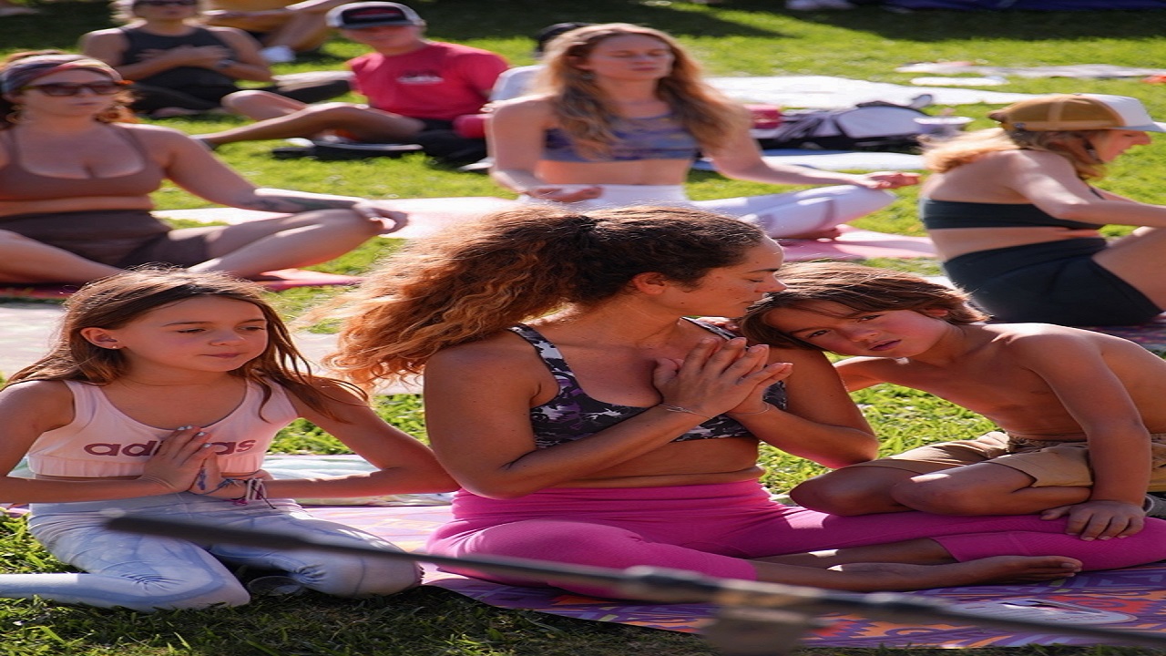 A woman and two children sit cross-legged on a mat outdoors, hands in prayer pose, with others practicing yoga on grass in the background. The scene is peaceful and sunlit. South Florida Business & Wealth