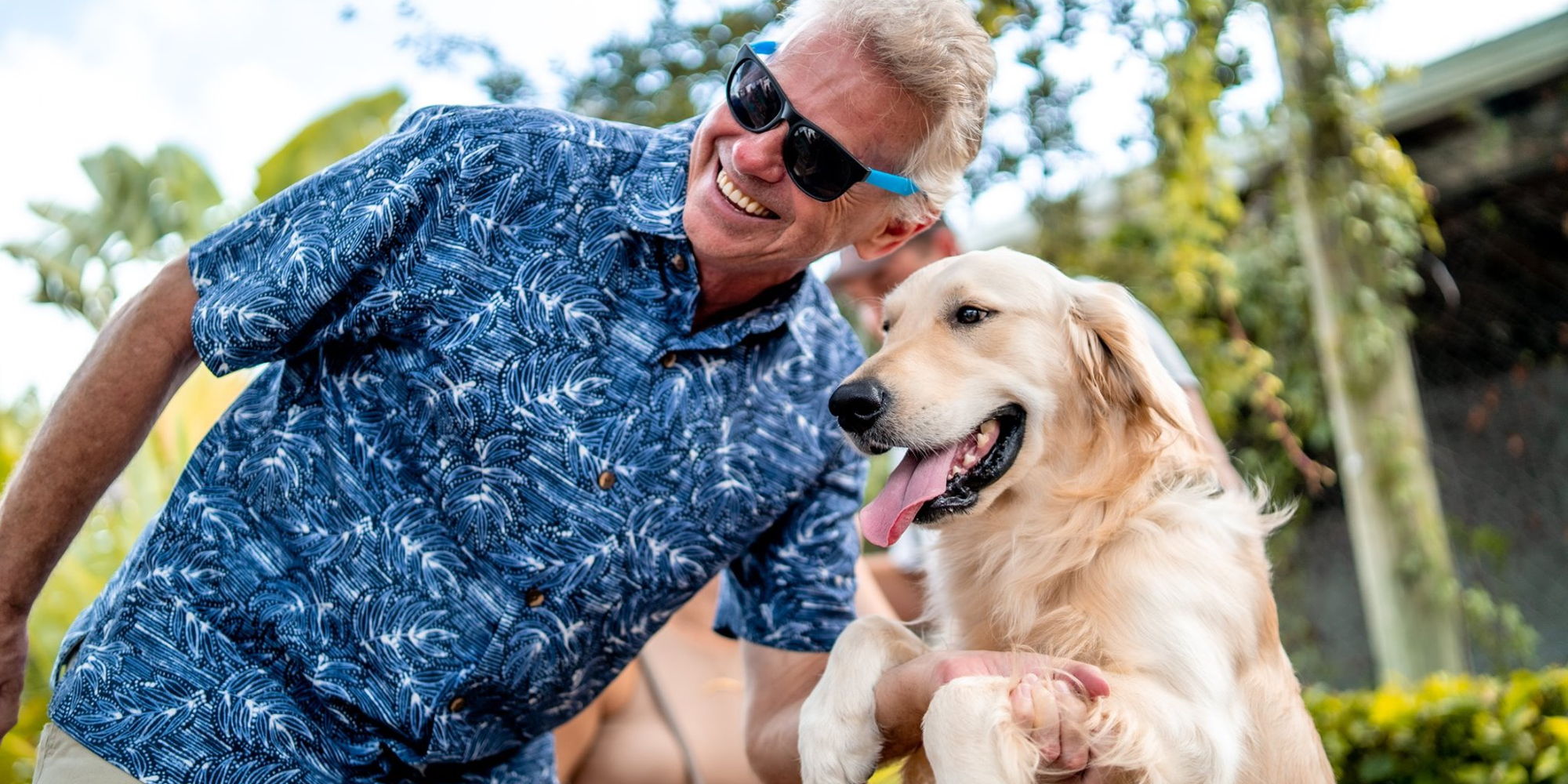 Smiling man in sunglasses and a blue patterned shirt kneels next to a happy golden retriever outdoors, both enjoying a sunny day. South Florida Business & Wealth