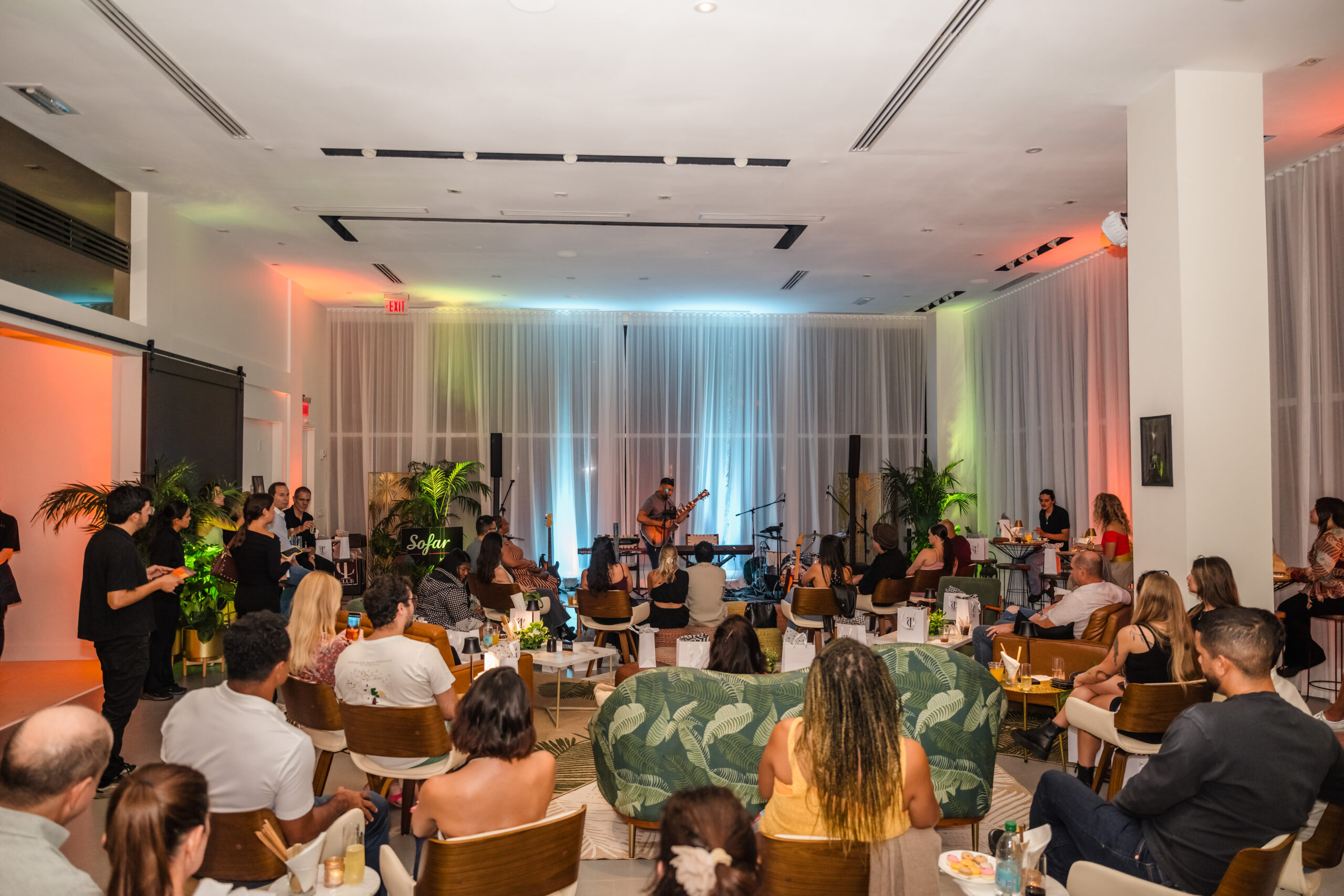 People sit in a modern, well-lit lounge watching a live band perform on stage. The room is decorated with plants, and soft colored lights illuminate the scene. Audience members face the musicians, enjoying the performance. South Florida Business & Wealth