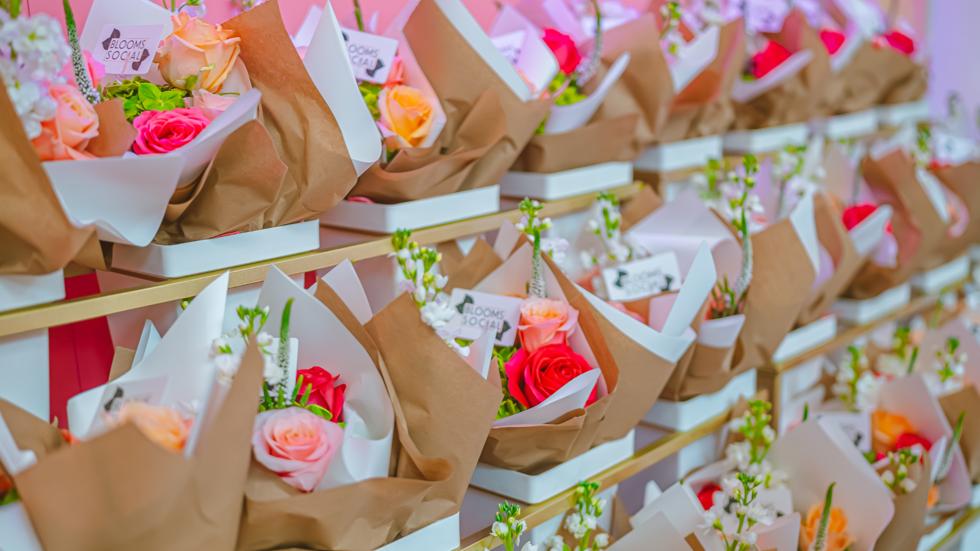 Multiple bouquets of colorful roses wrapped in white and brown paper are arranged on shelves, each with a small card tucked inside. There are pink, red, and orange roses with green leaves and white flowers. South Florida Business & Wealth