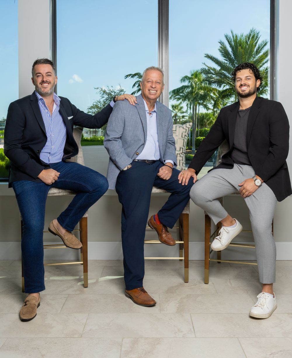 Three men wearing business casual outfits sit and stand near chairs in front of large windows with a tropical view and palm trees outside. They are all smiling and appear relaxed in a bright, modern room. South Florida Business & Wealth