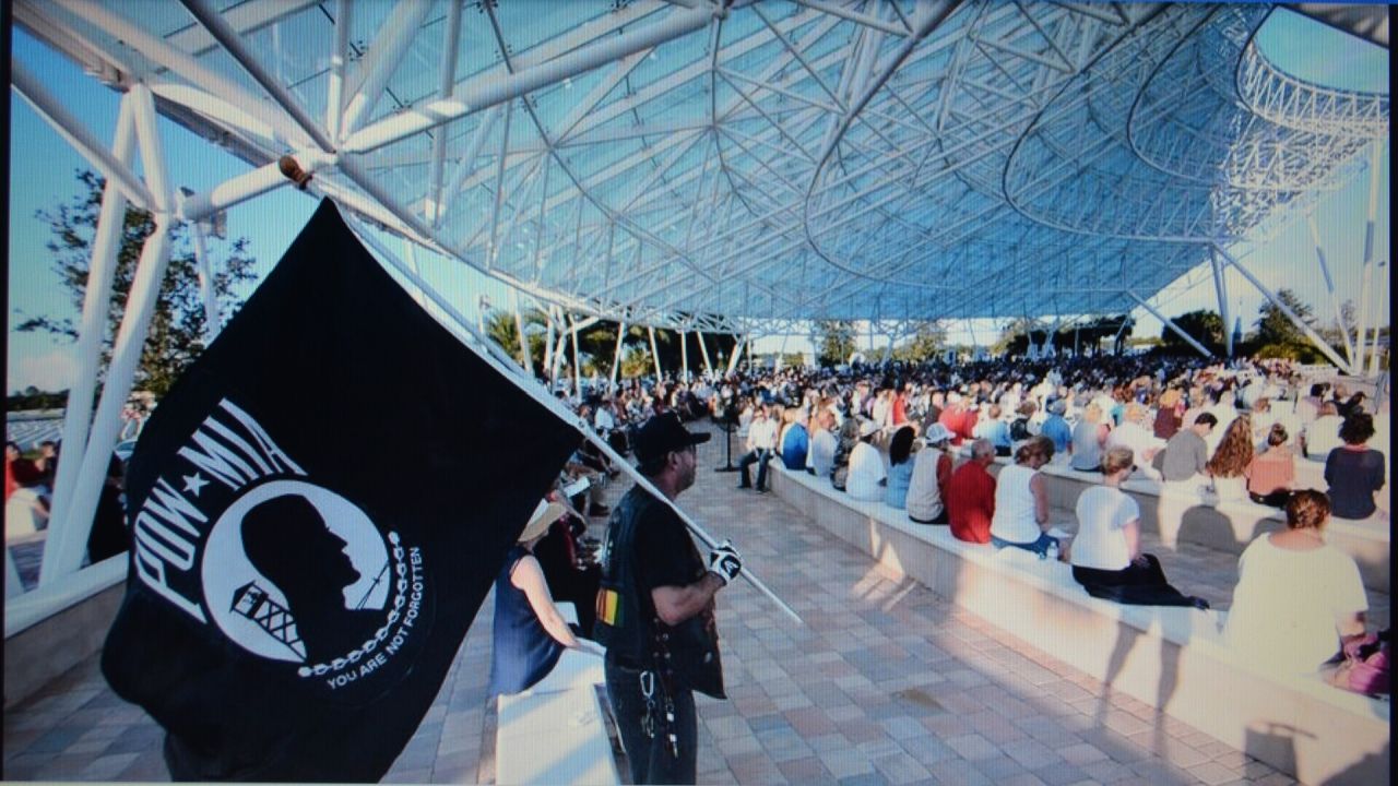 A person holding a POW-MIA flag stands in the foreground, while a large crowd sits under an open metal canopy at an outdoor event on a sunny day. South Florida Business & Wealth