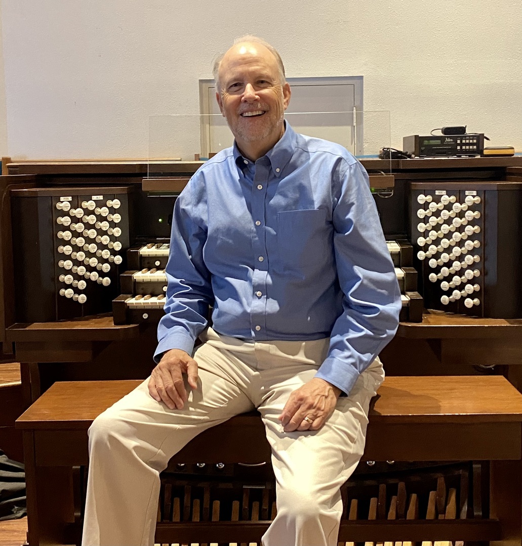 A man in a blue shirt and beige pants sits and smiles in front of a large pipe organ console with many buttons and keys. South Florida Business & Wealth