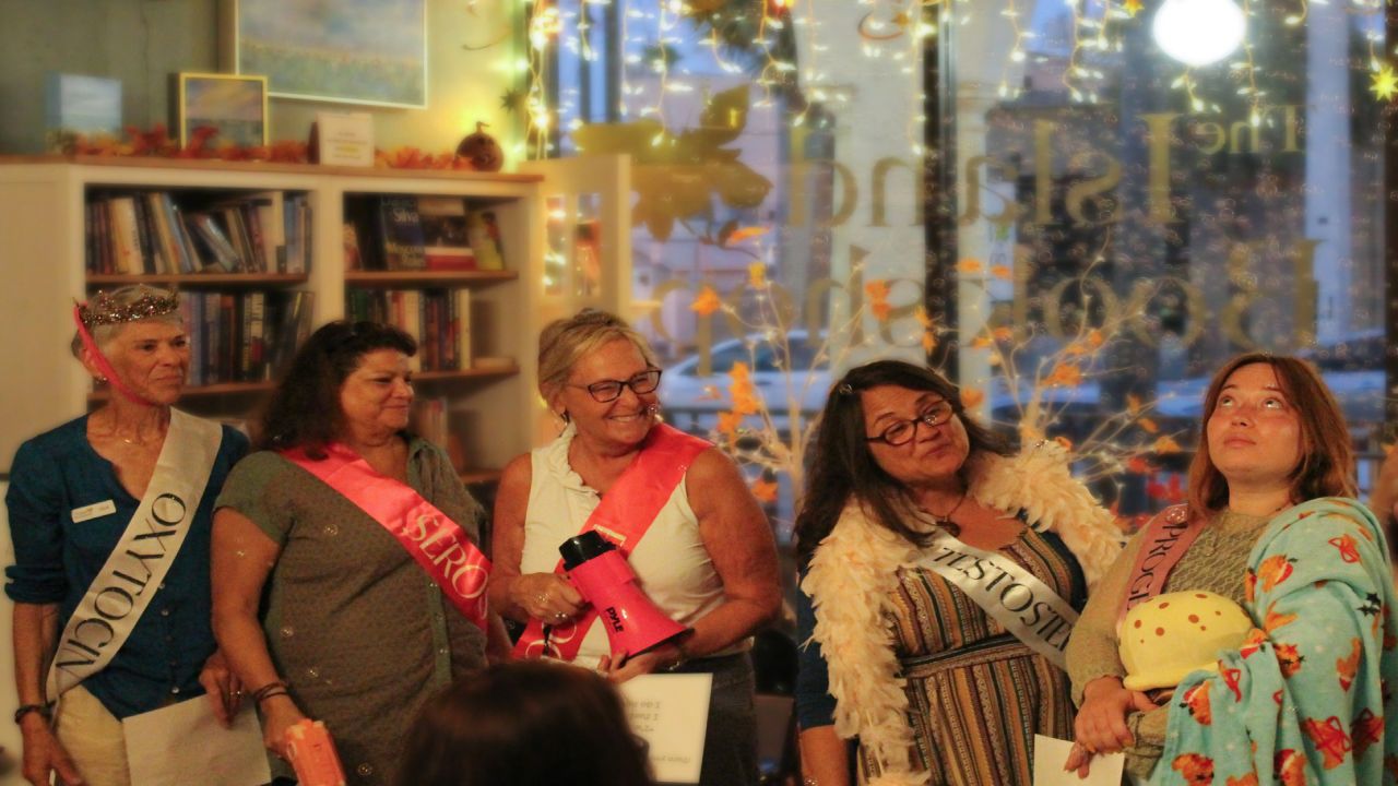 Five women wearing sashes and smiling stand together in a warmly lit, decorated room; one holds a crown, another a plush mushroom, with fairy lights and bookshelves in the background. South Florida Business & Wealth