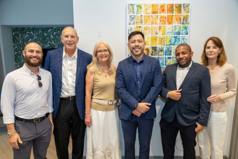 Six people stand indoors, dressed in business or business-casual attire, smiling at the camera. A colorful abstract artwork hangs on the wall behind them. South Florida Business & Wealth