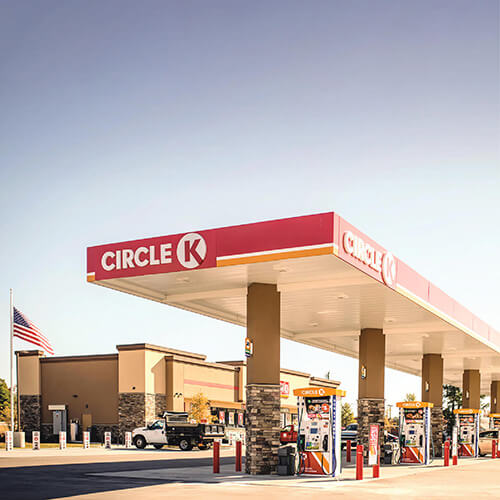 A Circle K gas station with fuel pumps under a red and white canopy, an American flag, and a convenience store building in the background on a clear day. South Florida Business & Wealth